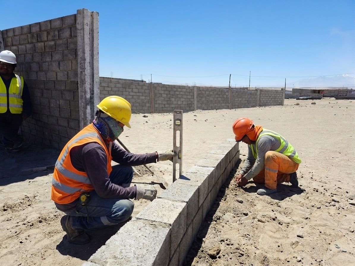 Dos operarios con casco y chaleco nivelan la base de un cerco junto a un muro terminado